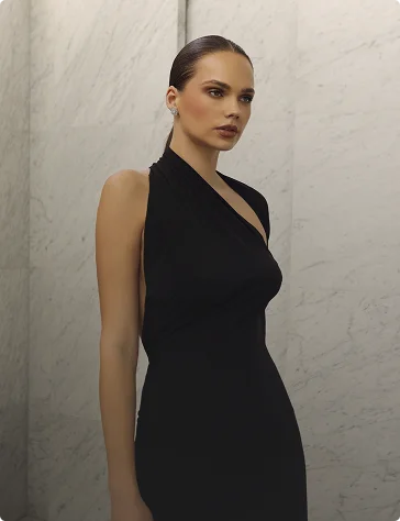 Model with slicked-back hair wearing a black one-shoulder dress against a marble wall.