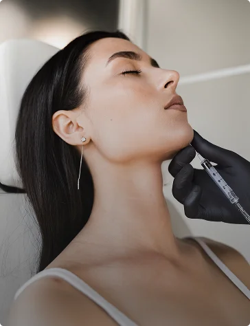 Side profile of a woman with dark hair receiving a chin injection from a clinician in black gloves.