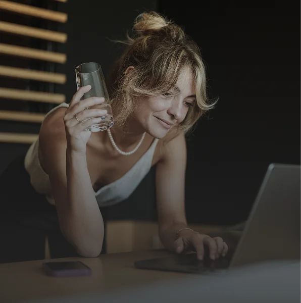 Woman in a white top leaning over a desk with a laptop and holding a glass of water.