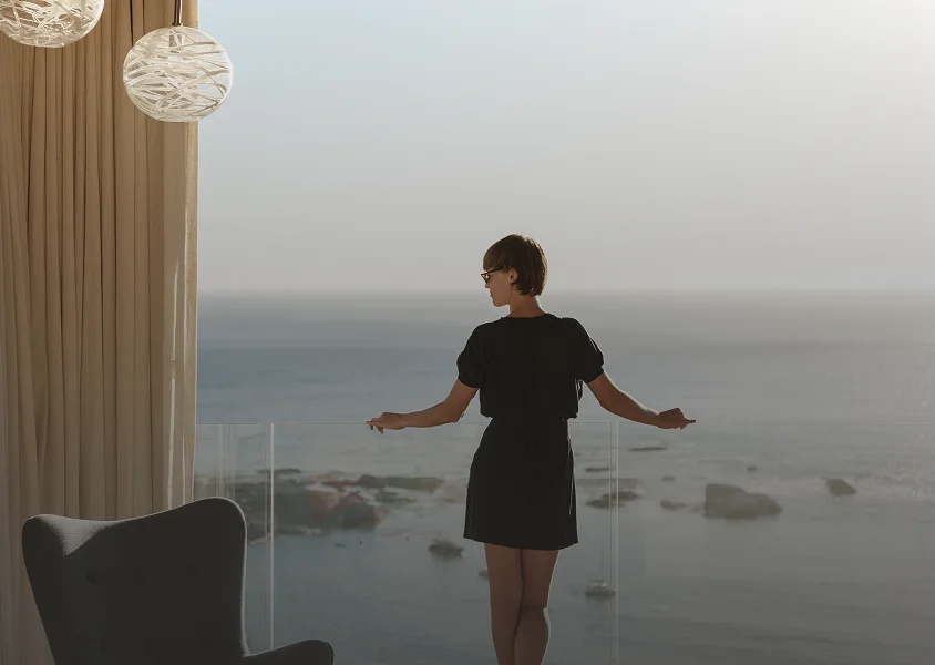 Woman in a black dress standing on a balcony overlooking the ocean at dusk.