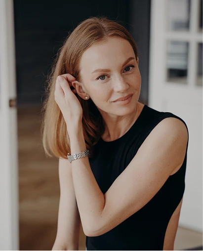 A woman with shoulder-length reddish-blonde hair and light freckles wears a black sleeveless top, a silver watch, and a stud earring, gently holding her hair back from her ear while looking at the camera against a grey background.