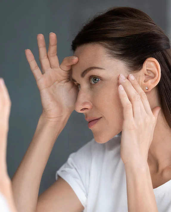 A middle-aged Caucasian woman is looking at her reflection in a mirror. She is touching the skin around her eyes with her fingers, pulling it taut, as if examining her eyelids and the area for signs of aging, like wrinkles or sagging skin. The woman has long, brown hair pulled back from her face. She is wearing a simple white t-shirt. The image is a close-up, focusing on her face and the act of self-examination. The woman's expression is serious and contemplative. - Laser Skin Resurfacing Fractional Ablative Non Ablative in Hermosa Beach, CA