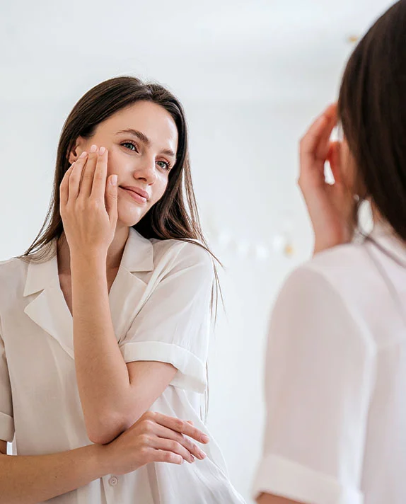 A woman in a white shirt looks into a mirror and gently touches her cheek with her hand. - Chemical Peels in Hermosa Beach, CA