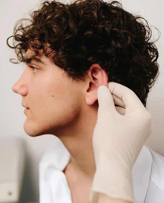 A profile view of a man with dark curly hair having his earlobe examined by a hand wearing a white sterile glove. - Earlobe Repair Piercing in Hermosa Beach, CA