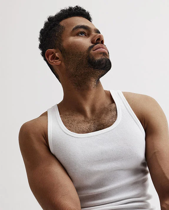A man with short dark curly hair and a beard wearing a white ribbed tank top looks upward against a plain light background. - Excessive Sweating in Hermosa Beach, CA