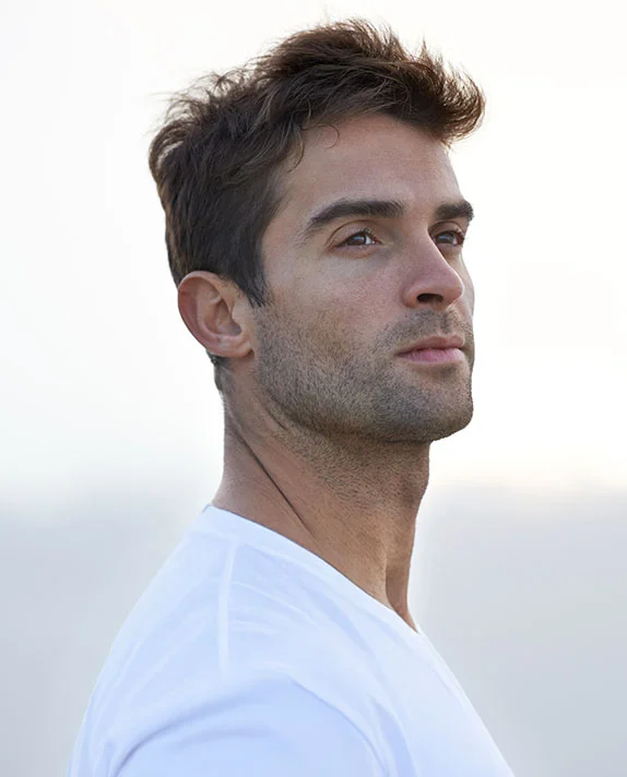 A man with short brown hair and stubble wearing a white t-shirt looks off into the distance. - Eyelid Heaviness in Hermosa Beach, CA