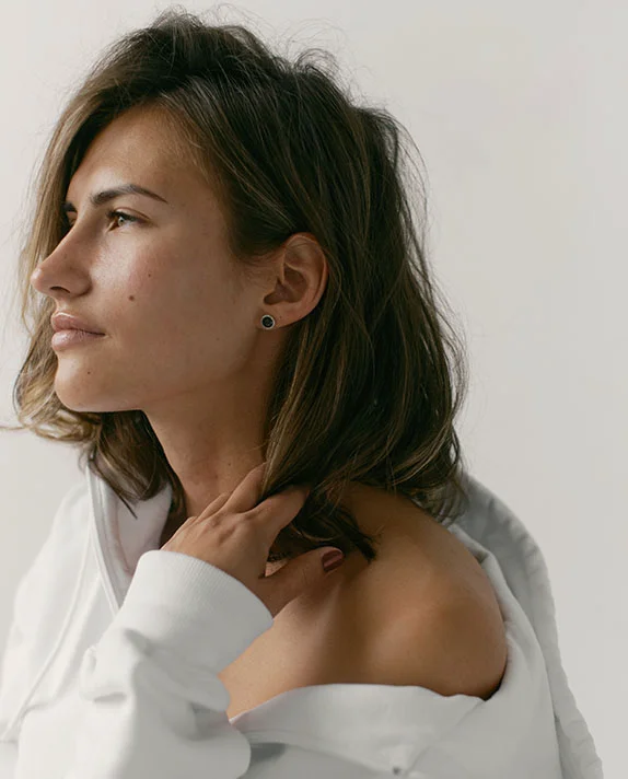 A young woman with brown, shoulder-length hair looks to the left, with her profile visible. She is wearing a white hoodie or top that is pulled down to expose her shoulder, and her hand is resting near her neck, gently touching her hair. She has a dark stud earring in her ear. The background is a plain, light color. - Fraxel Restore Dual in Hermosa Beach, CA