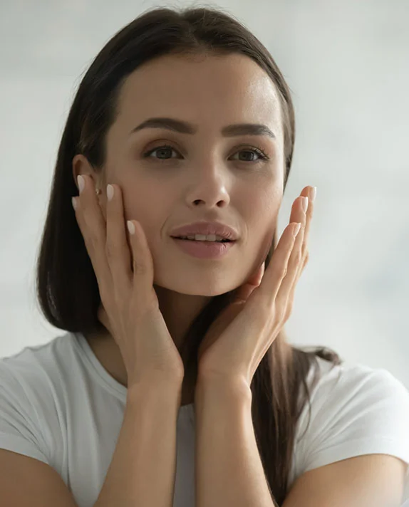 A young woman with long, dark hair looks directly at the camera, gently touching her cheeks with both hands. She is wearing a white t-shirt, and the background is blurred, showing a light-colored, possibly tiled, surface. - IPL in Hermosa Beach, CA