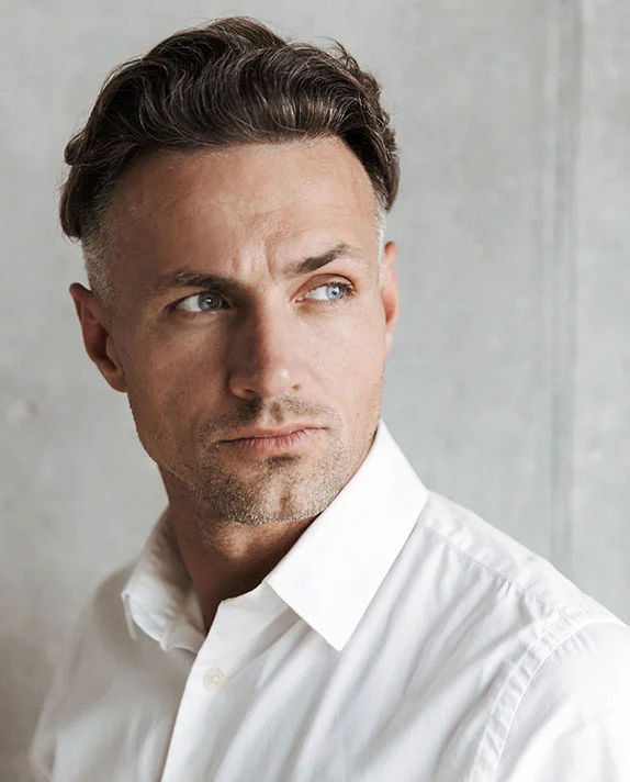 A man with dark and graying hair, wearing a white collared shirt, looking thoughtfully to his left. He has a serious expression on his face and is standing against a gray wall. - MOHs Surgery in Hermosa Beach, CA
