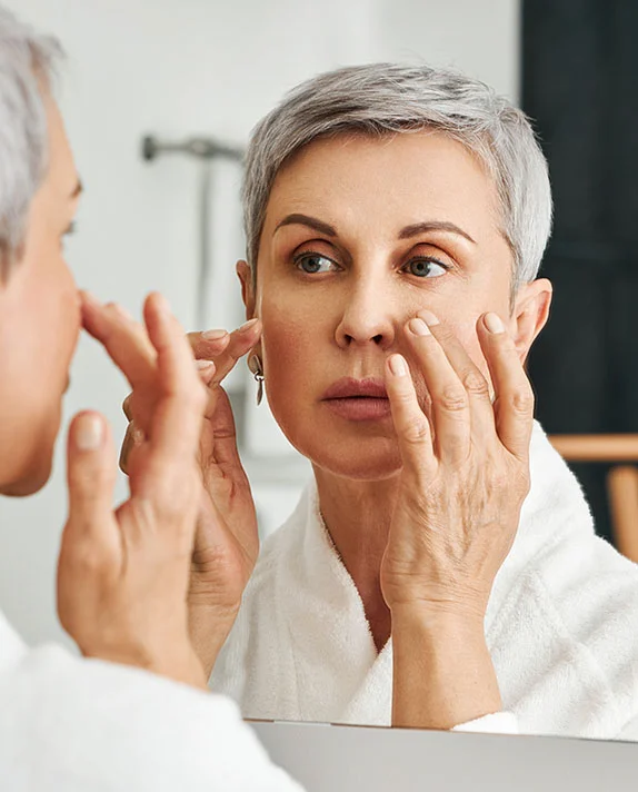 A woman with short, gray hair is looking at her reflection in a mirror. She is wearing a white bathrobe. She is touching her face, and her expression appears somewhat concerned or pensive. The lighting in the bathroom is bright and neutral. The background is a light gray/white bathroom wall. The reflection in the mirror shows a similar-aged woman with a similar expression. - Scar Revision in Hermosa Beach, CA